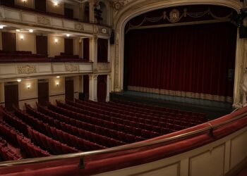 Leerer Theatersaal mit roten Sitzreihen und geschlossenem Bühnenvorhang, gesehen vom ersten Rang